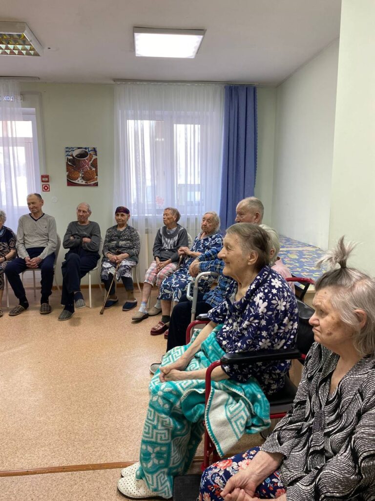 Группа пожилых людей сидит в зале полукругом